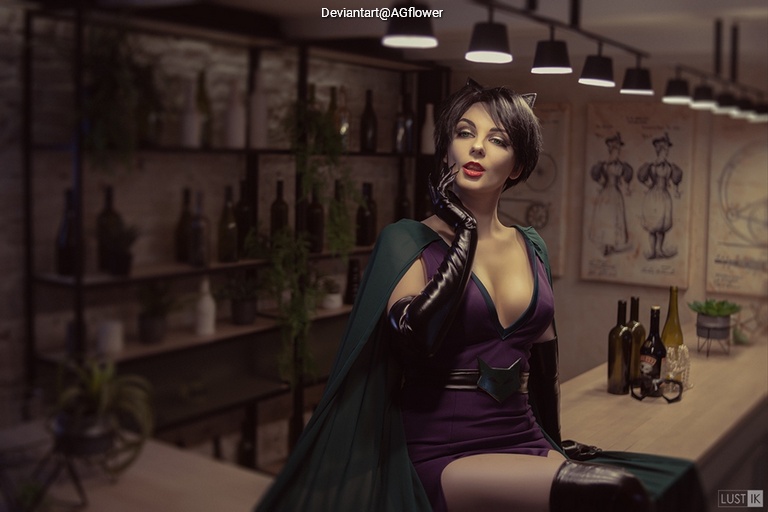 A woman in a purple dress and black gloves poses at a bar, wearing a green cape, with a stylish interior featuring bottles and art on the walls.