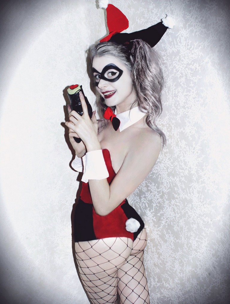 A woman dressed as a character with a red and black costume, featuring a jester's hat and fishnet stockings, holding a toy gun and smiling playfully.