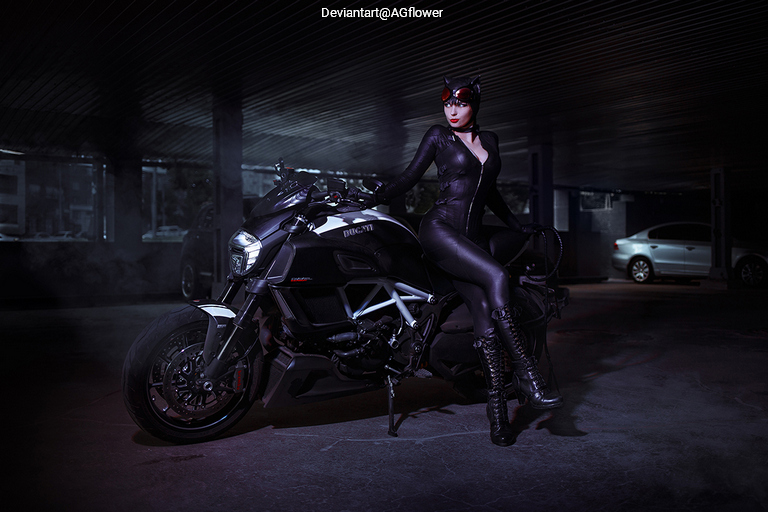 A woman dressed in a black cat-themed costume posing next to a sleek motorcycle in a dimly lit parking garage.