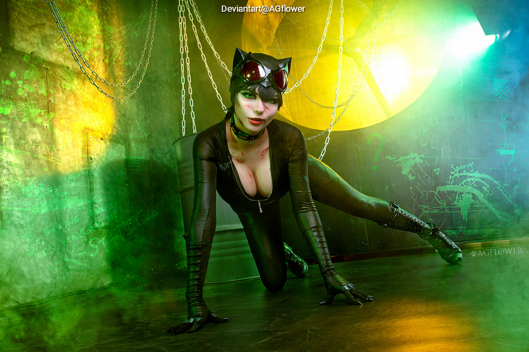 A woman in a black catsuit with cat ears and goggles poses in a dramatic, colorful setting with chains and mist.