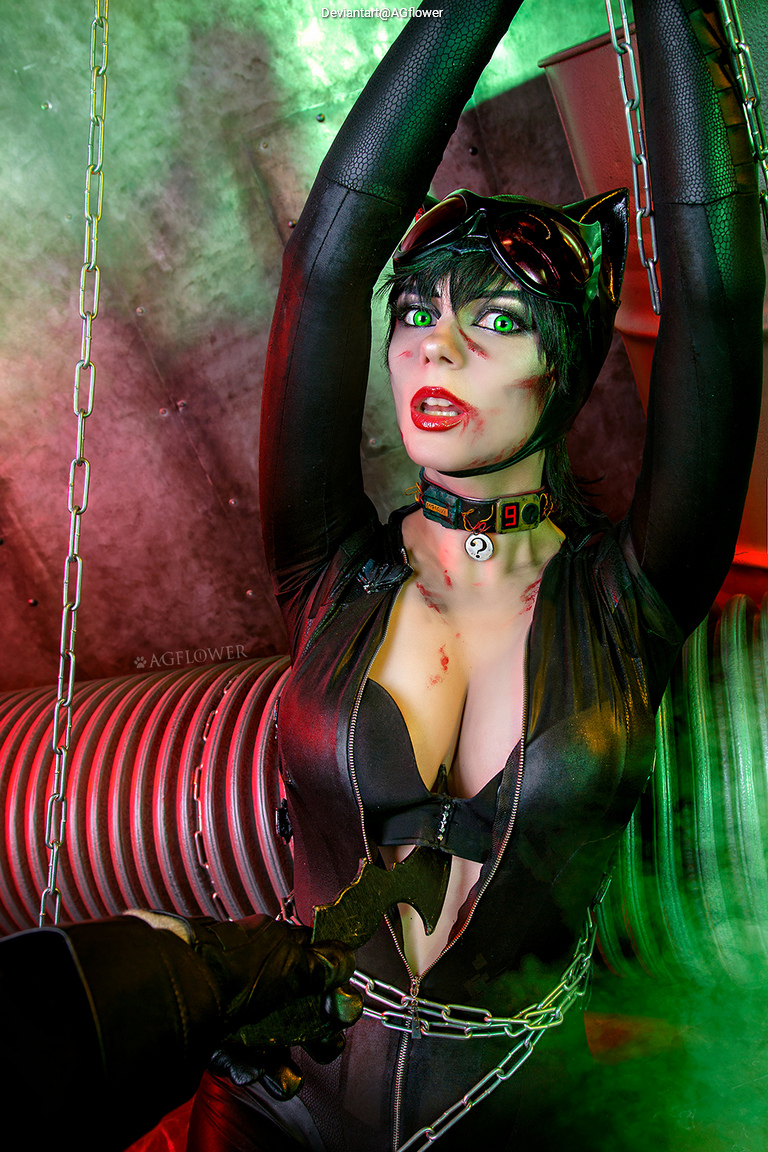 A female cosplayer dressed as Catwoman, with a black leather outfit and cat ears, is being held captive with chains. She has a frightened expression, green eye makeup, and visible scratches on her face. A hand in the foreground holds a weapon, suggesting a tense confrontation.