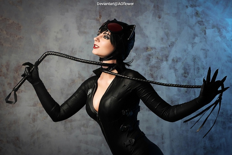 A woman dressed as a cat-themed character, wearing a black outfit and cat ears, playfully holding a whip against a textured backdrop.