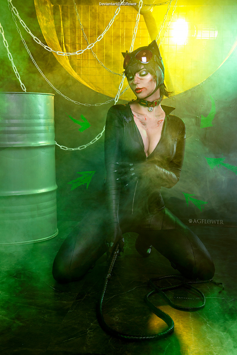 A person dressed as a cat burglar, kneeling on the floor in a smoky, green-lit environment with chains and barrels in the background, holding a whip.
