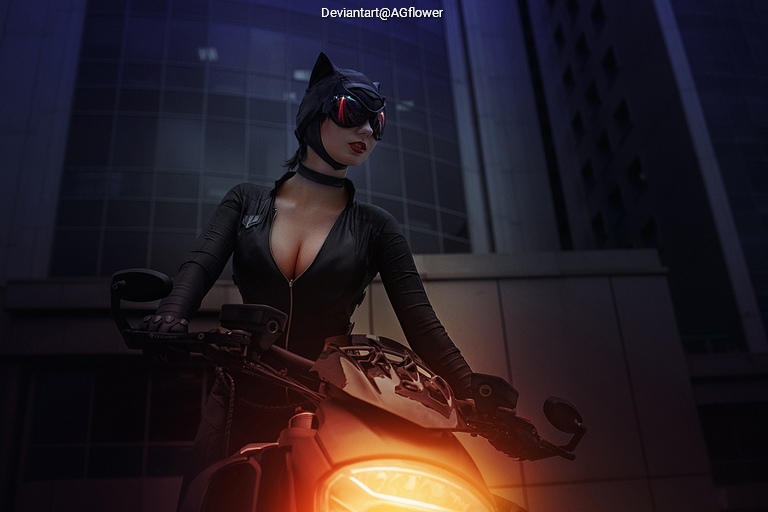 A woman in a black catsuit and helmet poses confidently on a motorcycle in an urban setting at night.