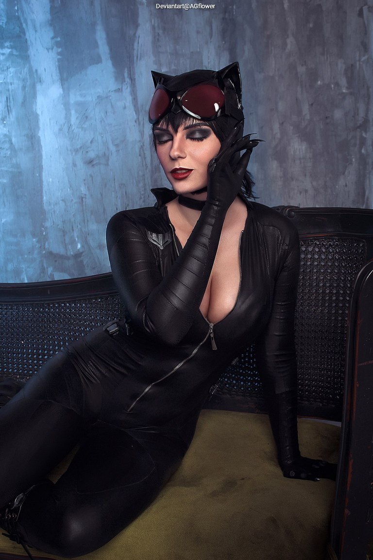 A woman in a black cat-themed costume poses elegantly on a couch, wearing gloves and goggles, with a dramatic makeup look.