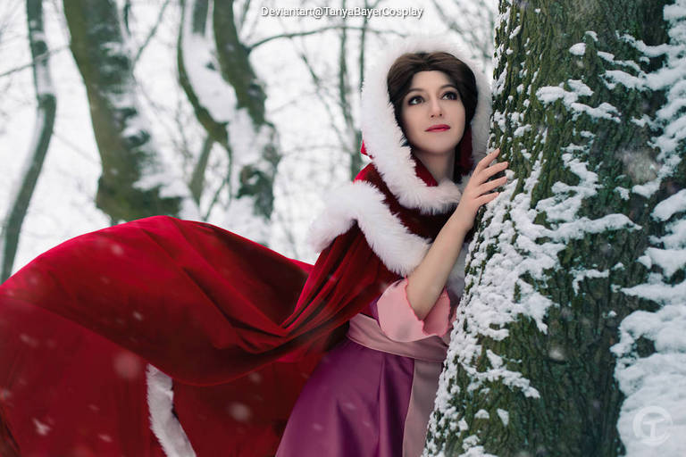 A cosplayer dressed in a red cloak and pink gown stands next to a snow-covered tree, gazing thoughtfully into the distance.