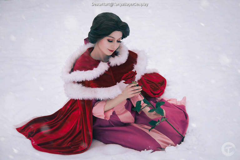 A woman in a red and white fur-trimmed cape, sitting in the snow, delicately holding a red rose.