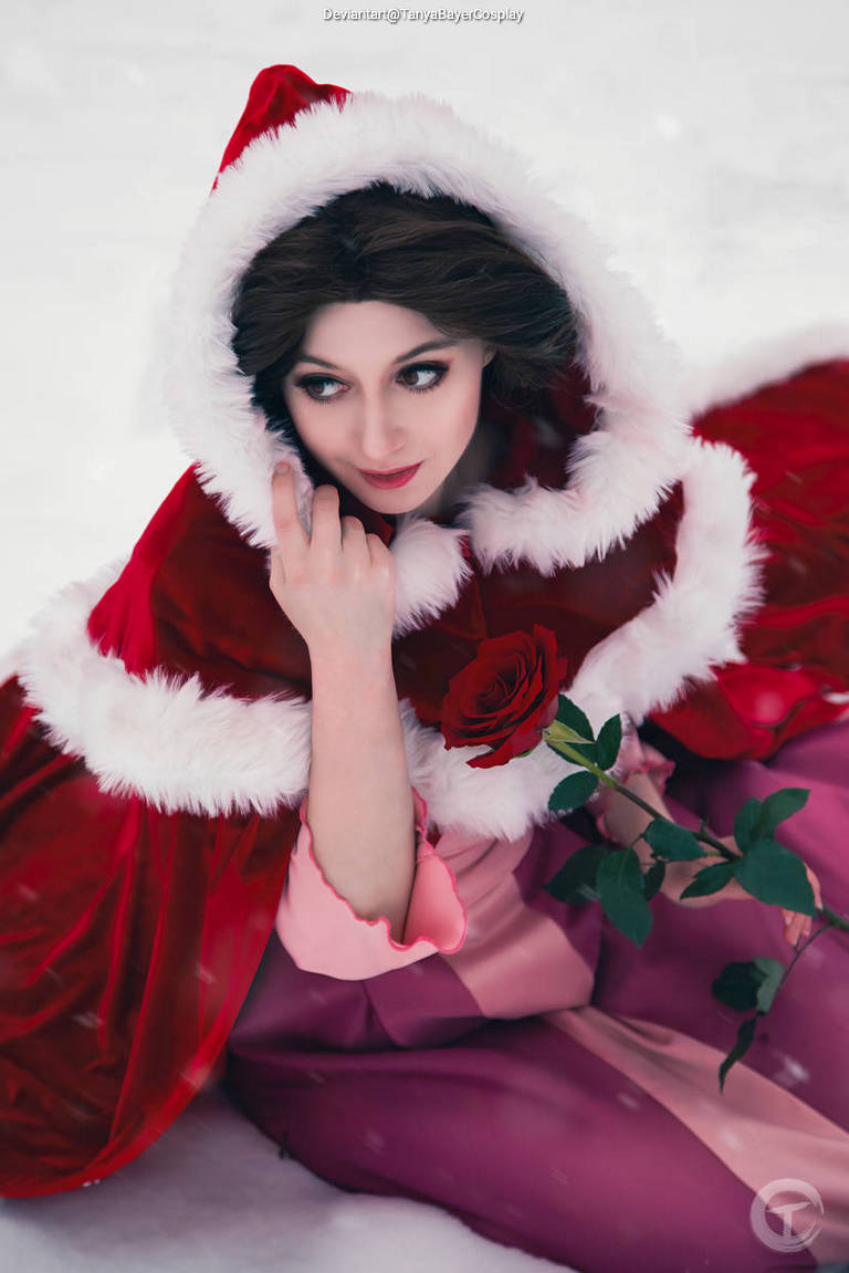 A person dressed in a red and white hooded cloak holds a rose while sitting in a snowy landscape, with a soft expression on their face.