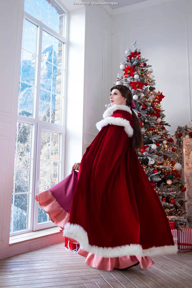 A woman in a red cape with white fur trim stands gracefully near a decorated Christmas tree, with snowy mountains visible through large windows in the background.
