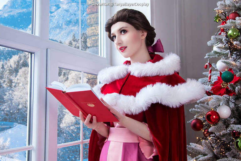 A woman in an elegant red cape with white fur trim, holding a book, stands by a snowy window, with a decorated Christmas tree nearby.