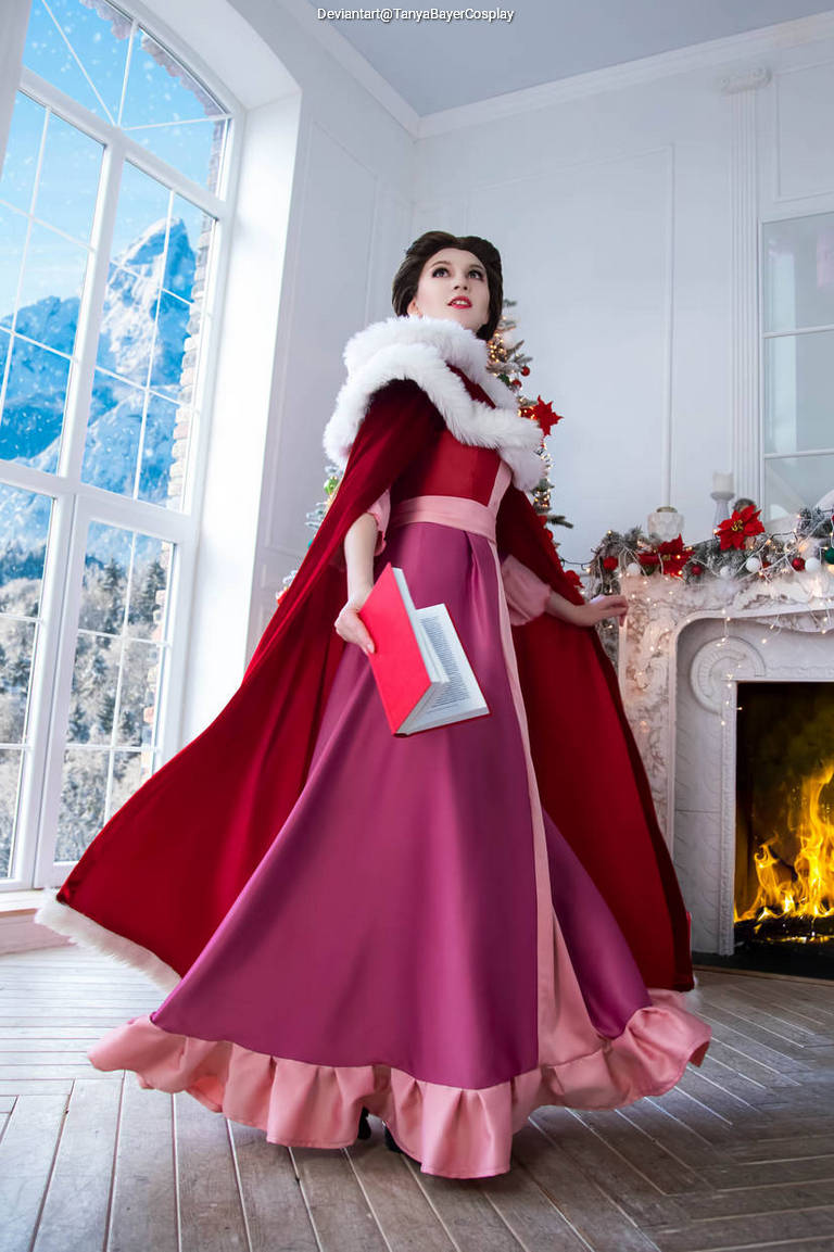 A woman in a pink and red gown with a white fur collar stands elegantly in a beautifully decorated room, holding a book. The background features snowy mountains and a cozy fireplace.