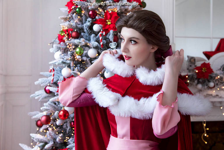 A woman in a red and pink costume with a fur trim adjusts her hair, standing in front of a decorated Christmas tree.
