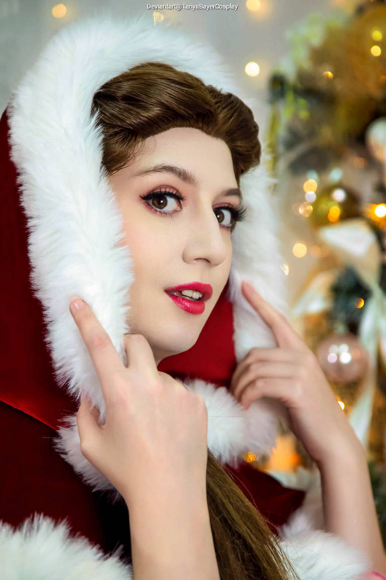 Close-up of a person in a red hooded cloak with white fur trim, posing with fingers near their face, set against a festive decorated background.