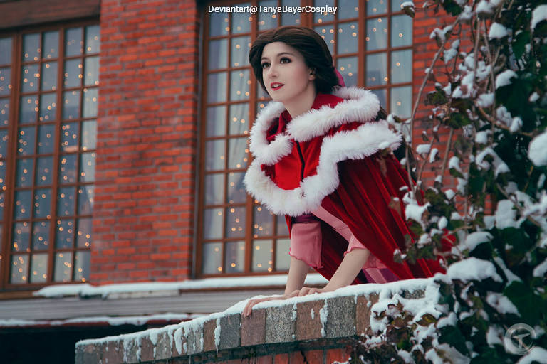 A woman in a winter outfit with a red cloak and white fur trim gazes out thoughtfully from a snowy ledge, with twinkling lights in the background.