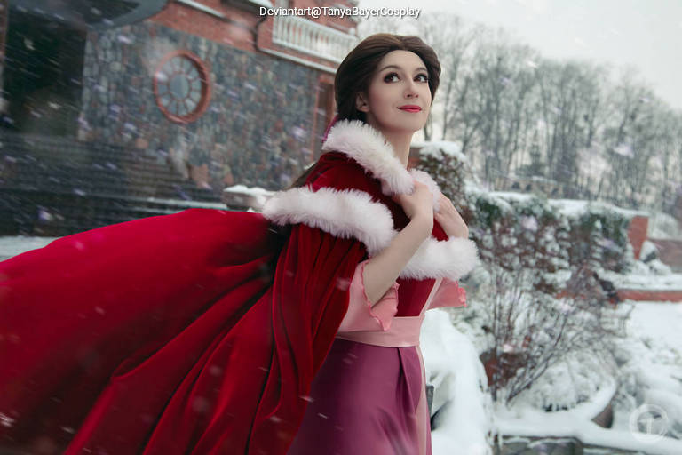 A woman in an elegant red cape with white fur trim stands in a snowy landscape, looking thoughtfully into the distance.