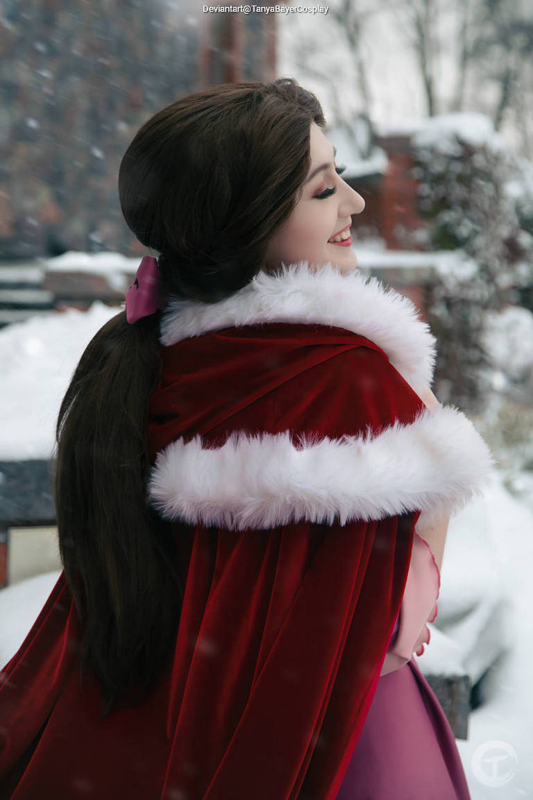 A young woman with long dark hair, wearing a red velvet cape with white fur trim, smiles while standing in a snowy outdoor setting.