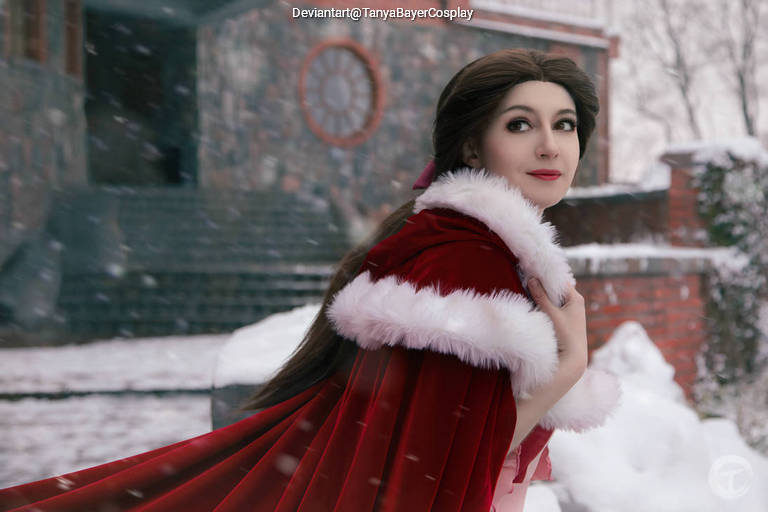 A woman with long brown hair wearing a red cape with white fur trim poses in a snowy outdoor setting, with a stone building in the background.