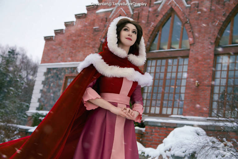 A woman cosplaying in a winter-themed outfit, featuring a red cloak with white fur trim and a long pink dress. She stands outside in the snow, in front of a brick building with large windows.