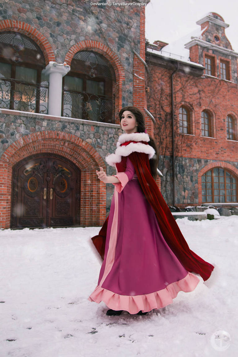 A woman in a pink dress with a red cape stands in the snow, smiling softly. She is in front of a brick building with large wooden doors.