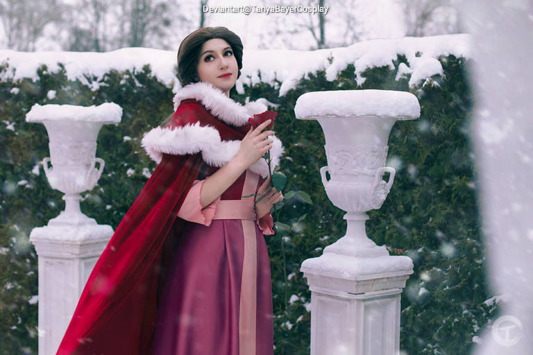A person in a winter setting wearing a red cape with white fur trim, holding a red rose, standing near white decorative vases surrounded by snow-covered greenery.