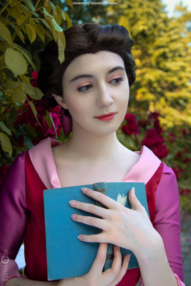 A person in a pink and purple outfit holding a blue book, surrounded by greenery and flowers.