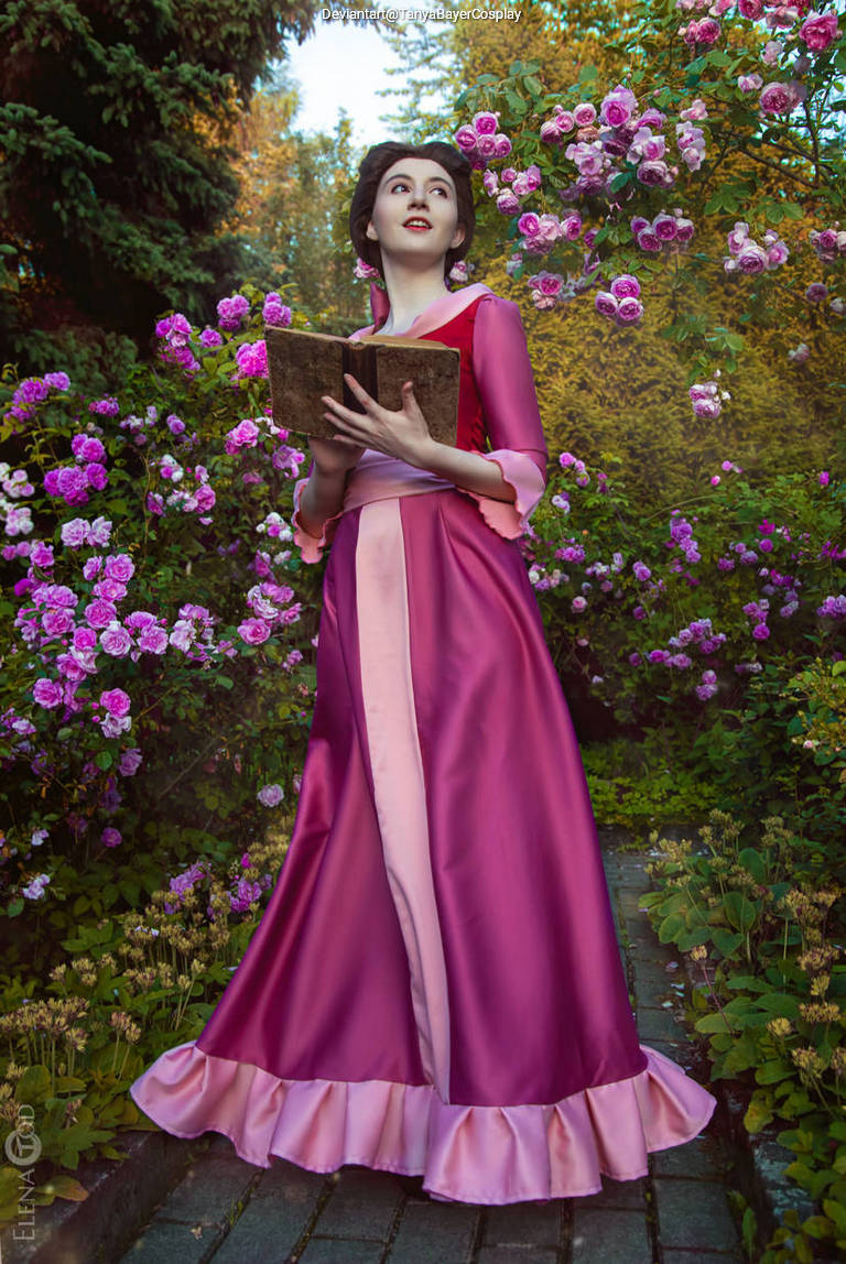A person in a pink dress, holding a book, standing amidst blooming flowers in a garden.