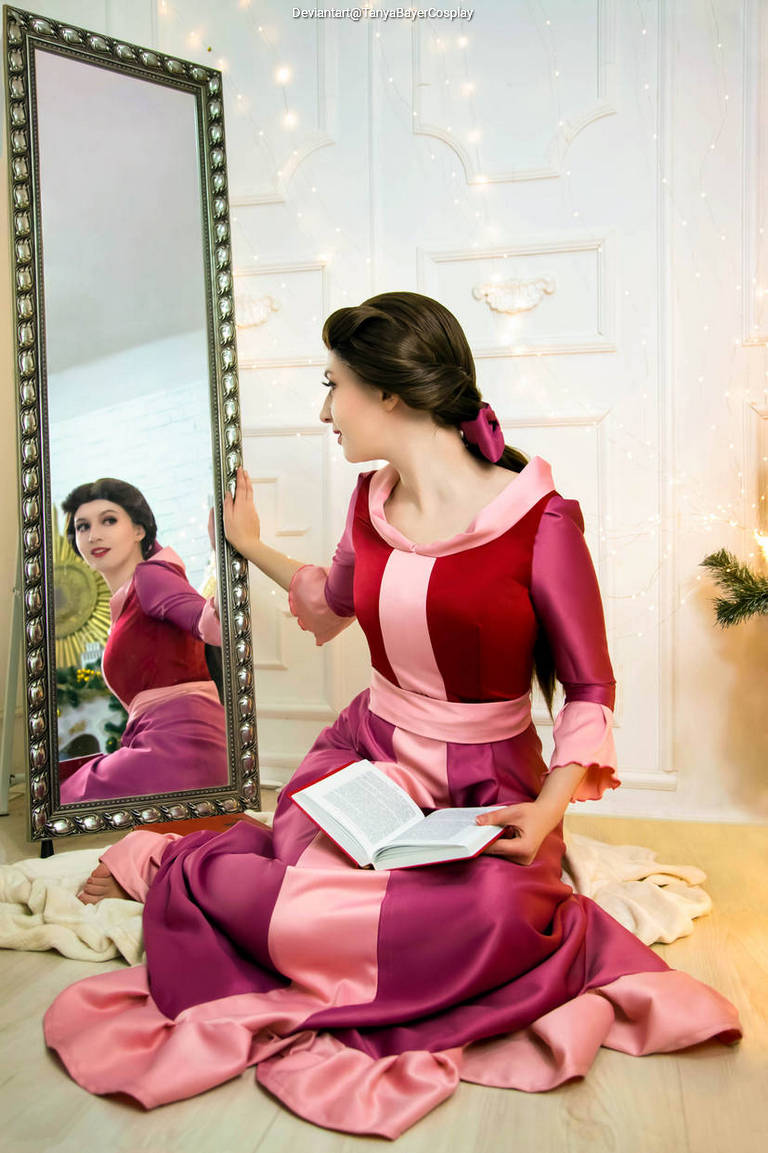 A woman in a pink and red dress sits on the floor, looking at her reflection in a large mirror while holding a book.