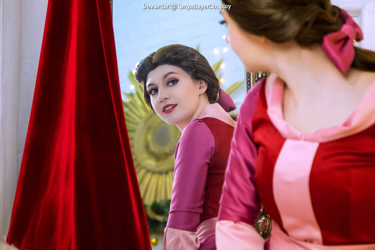 A cosplayer dressed in pink and red outfits looks back at her reflection in a mirror, with a graceful smile, surrounded by warm lighting and a decorative background.