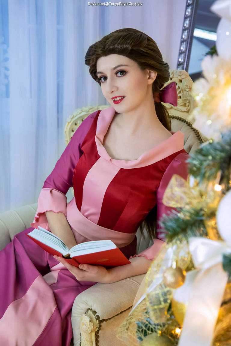 A person in a pink and red dress sitting on a chair, holding a book, with a decorated Christmas tree in the background.