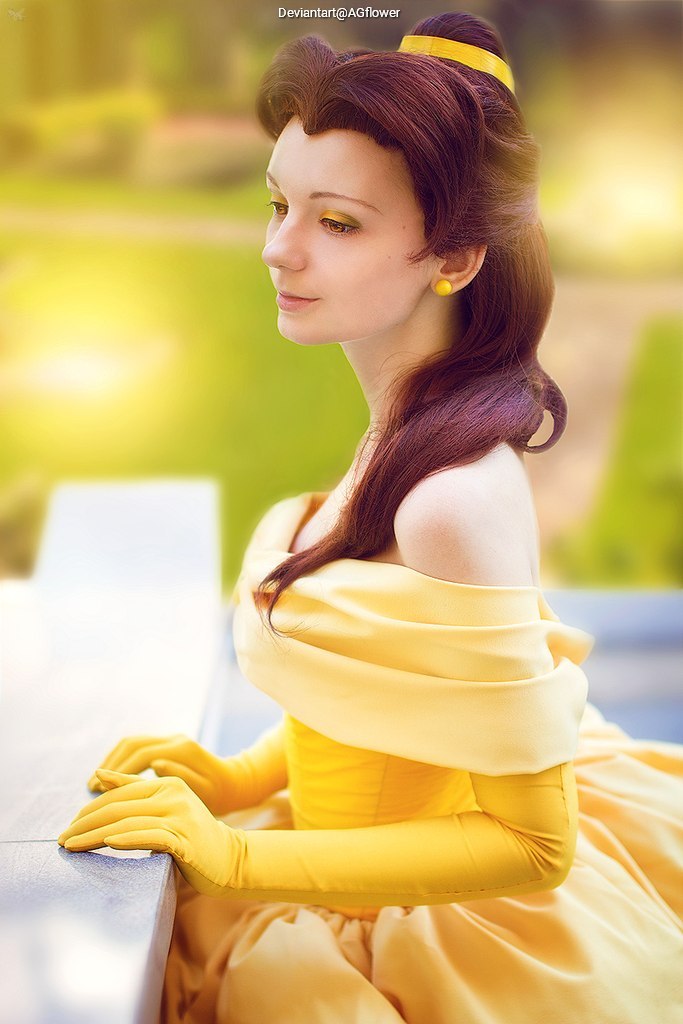 A woman in a yellow ball gown with matching gloves and hair accessories, seated outdoors with a serene expression.