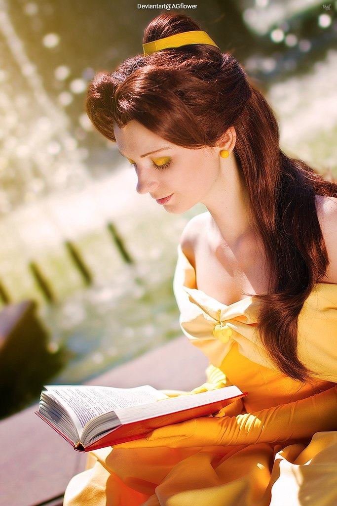 A woman dressed as a princess with brown hair styled in an elegant updo, wearing a yellow dress and yellow earrings, is sitting outdoors reading a book, with soft sunlight and a blurred fountain in the background.