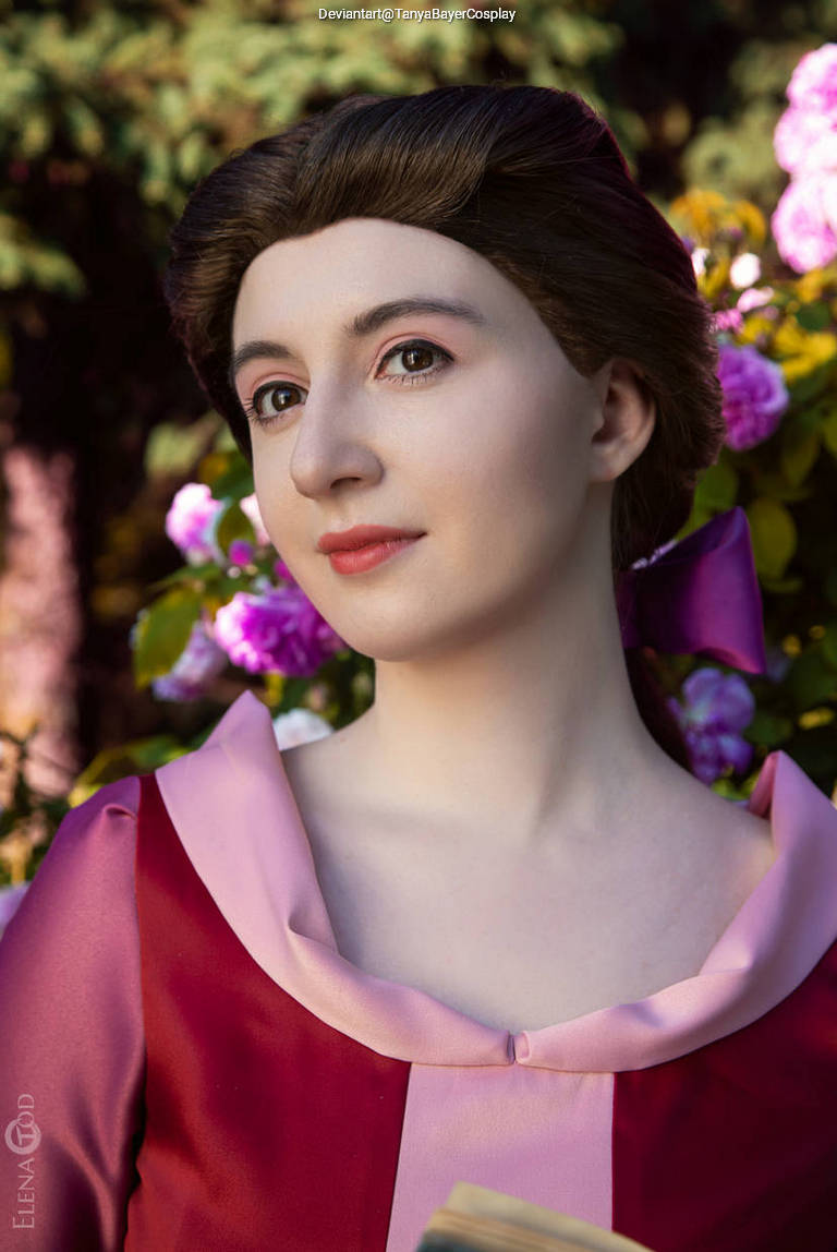 A person in a pink and red dress, standing among flowers, looking thoughtfully at the camera.
