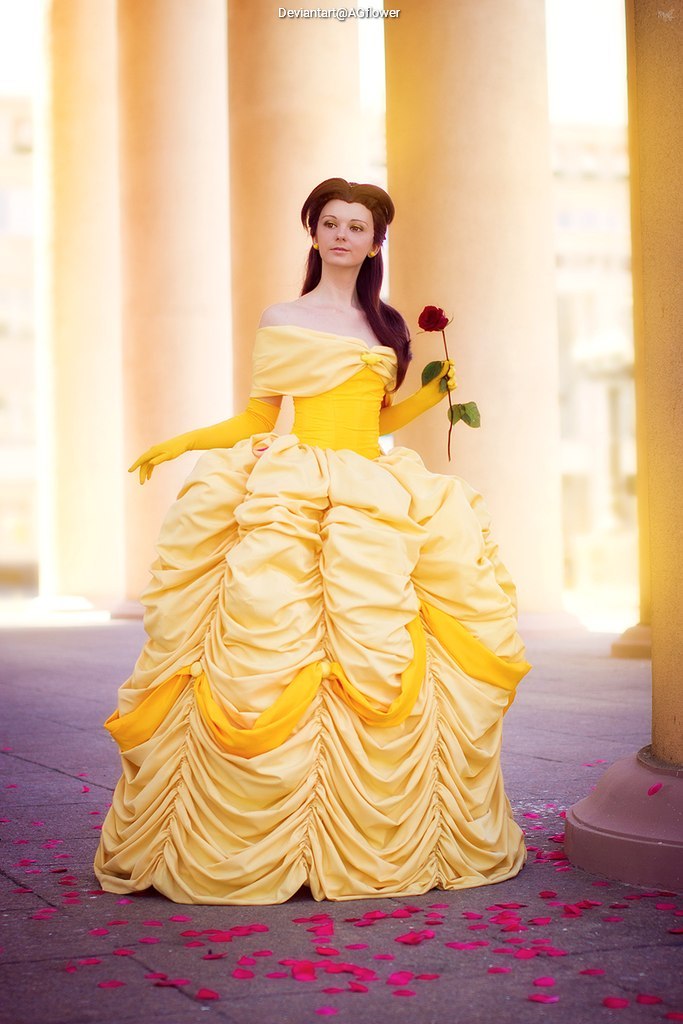 A person dressed in a yellow gown inspired by a fairy tale character, holding a red rose, standing among scattered rose petals against a backdrop of columns.