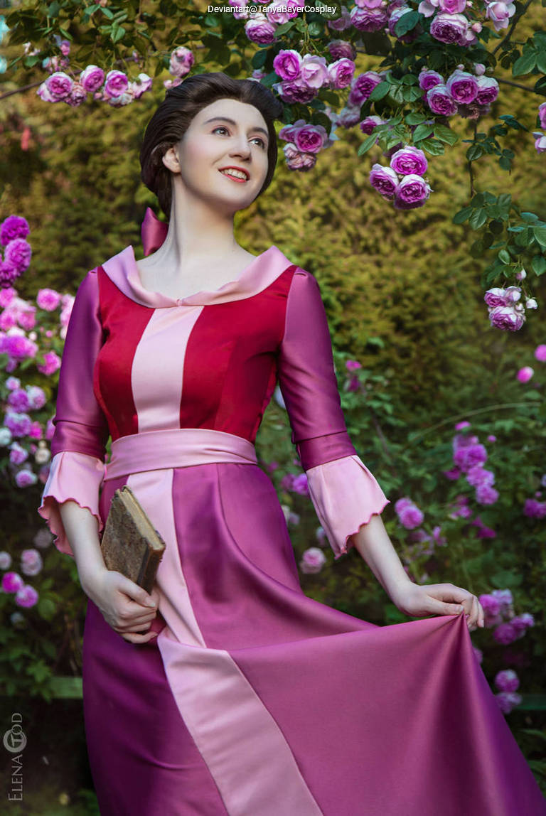 A woman wearing a colorful pink and red dress with long sleeves stands in a garden surrounded by blooming pink roses, smiling and holding a book in one hand.