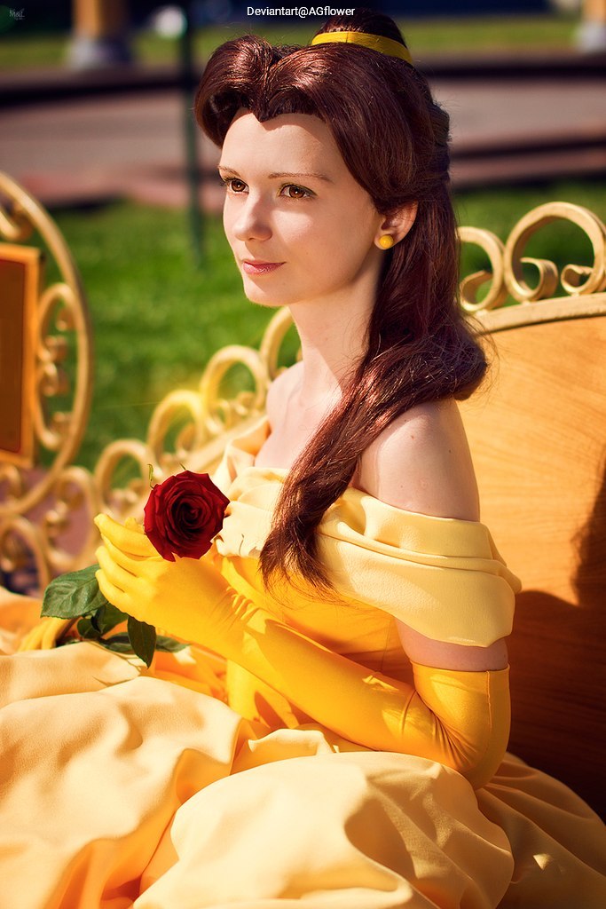 A woman dressed as a character in a golden yellow gown, holding a red rose, seated on a wooden bench in a sunny outdoor setting.