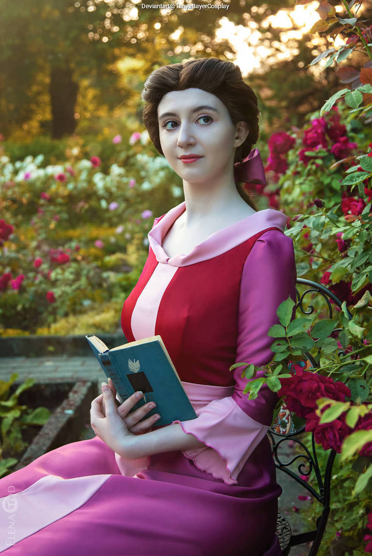 A woman dressed in a pink and red gown sits on a garden bench, holding a closed book and gazing away, surrounded by colorful flowers.