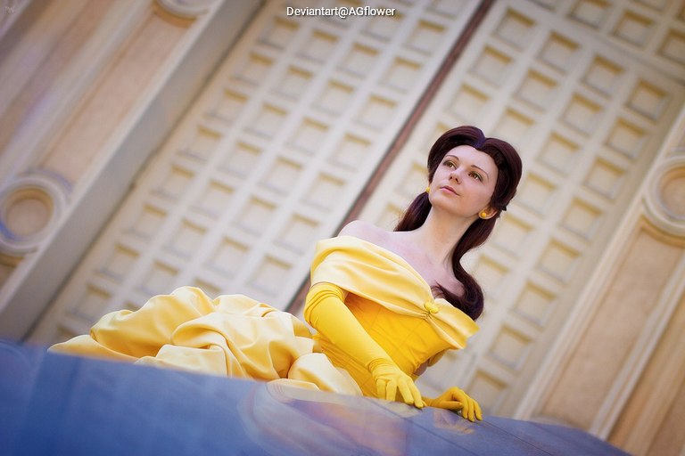 A woman dressed as Belle from 'Beauty and the Beast,' wearing a yellow gown and gloves, posed thoughtfully in front of a textured backdrop.