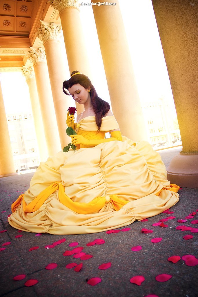 A woman in a yellow gown reminiscent of a fairy tale character sits gracefully on the ground, holding a red rose, with rose petals scattered around her.