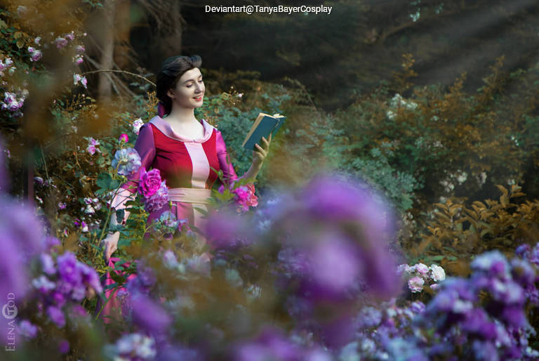 A woman in a pink and purple dress stands in a garden filled with colorful flowers, smiling while holding an open book.