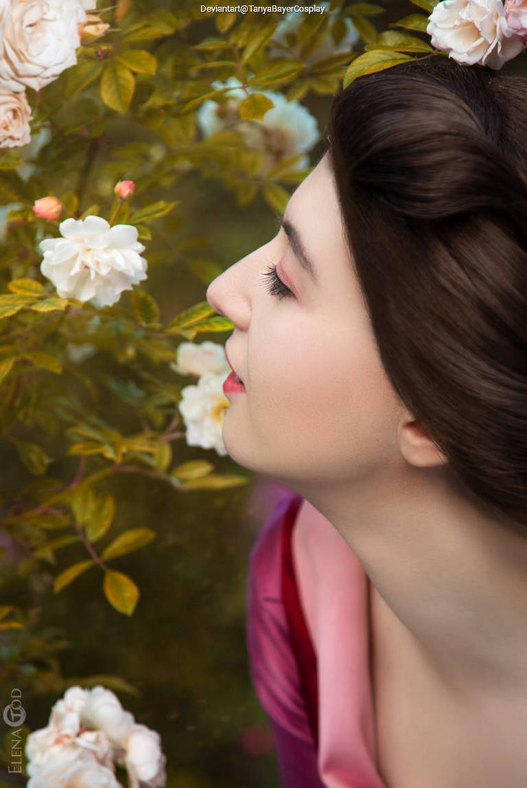 A woman with dark hair and soft makeup gazes thoughtfully at blooming roses in a garden, wearing a pink dress.