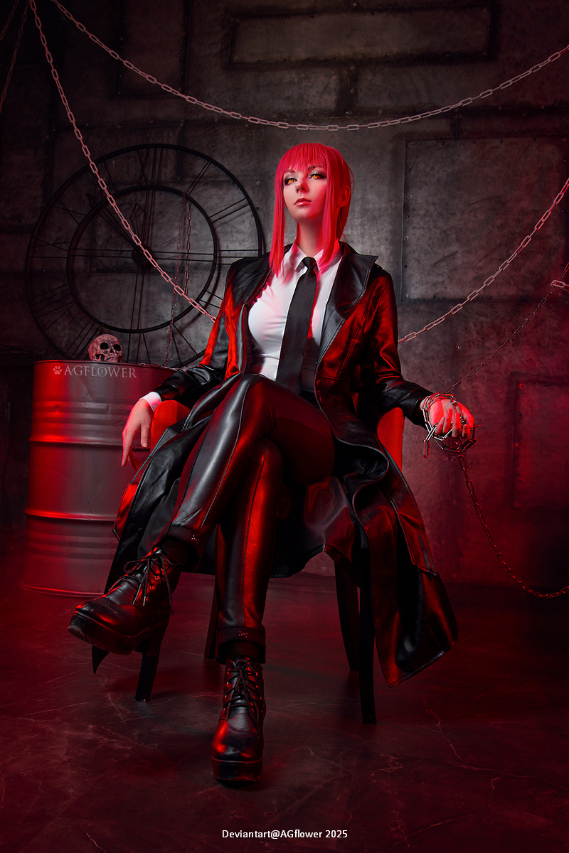 A woman with red hair is sitting in a dark, industrial setting, wearing a black suit and tie, with a long coat. The scene is illuminated in red light, and she is holding chains in her hands while sitting confidently in a chair.