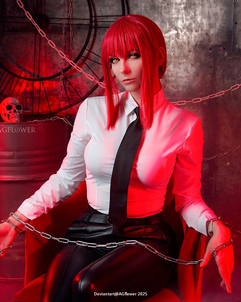 A person with red hair and striking eyes dressed in a white shirt and black tie, sitting in a chair with chains around their wrists, against a backdrop of red lighting and metallic elements.