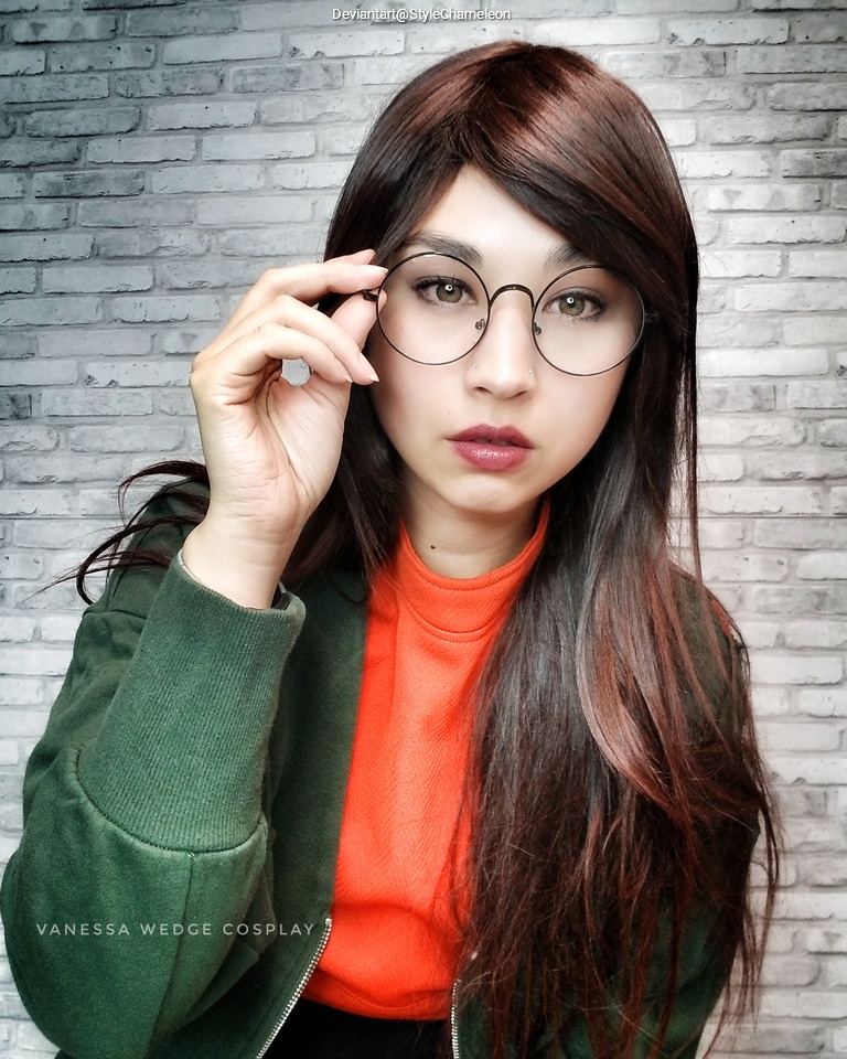 A woman with long brown hair, wearing round glasses and an orange turtleneck under a green jacket, posing in front of a brick wall.