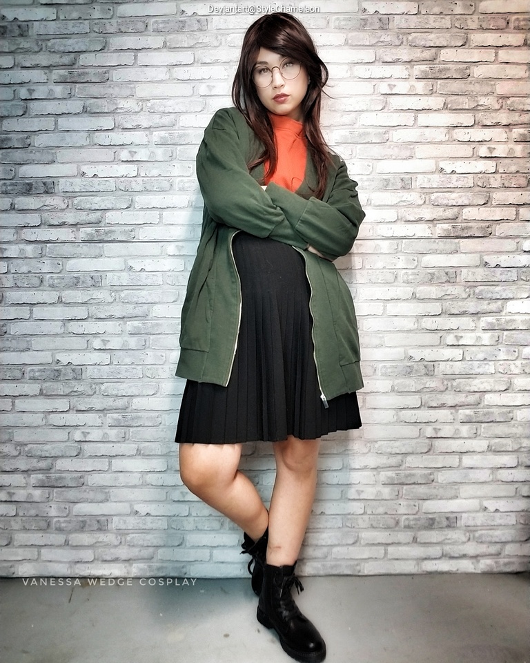 A person standing in front of a brick wall, wearing a green jacket over an orange top and a black pleated skirt, with glasses and black boots.