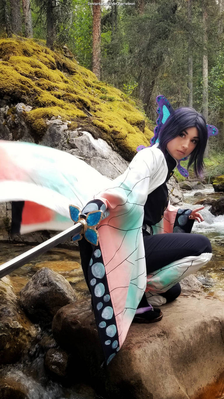 Person in colorful traditional attire with butterfly-themed details, crouching on a rock near a stream, holding a sword.