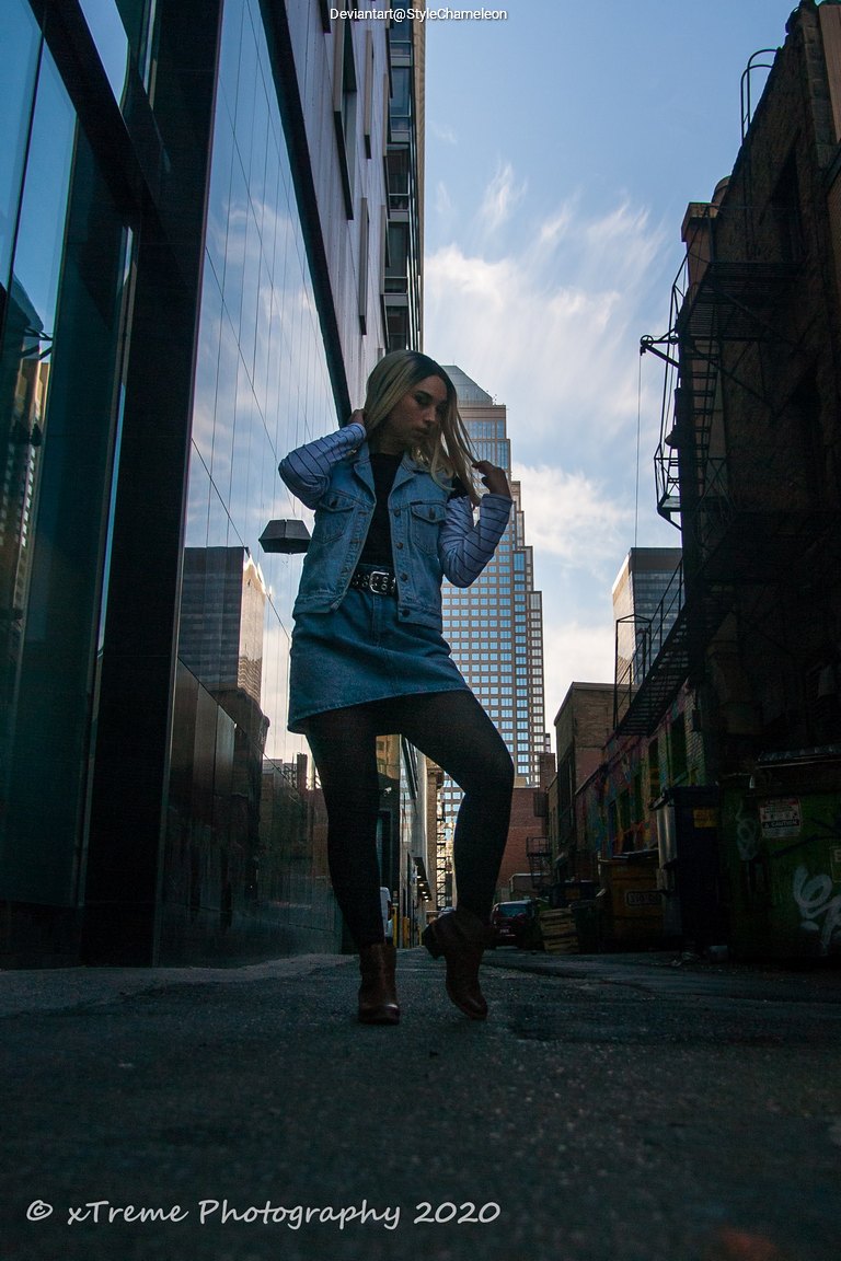 A person with long blonde hair wearing a denim jacket and skirt is posing in an urban alleyway, with buildings and a blue sky in the background.