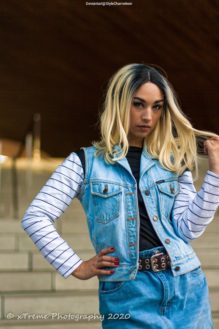 A person with long blonde hair styled in waves, wearing a denim vest over a black shirt and striped long-sleeve top, posing confidently with one hand in their hair and the other on their hip. The background features a staircase.