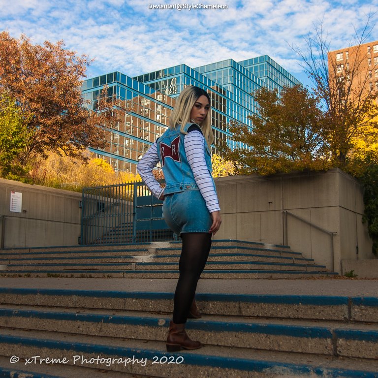A young woman with long, light-colored hair wearing a denim outfit with a red emblem, standing on stairs in an urban setting with modern buildings and autumn foliage in the background.