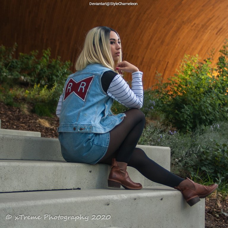 A person sitting on concrete steps, wearing a denim vest with a red 'RR' emblem and a striped long-sleeve shirt, paired with tights and brown ankle boots, surrounded by greenery.