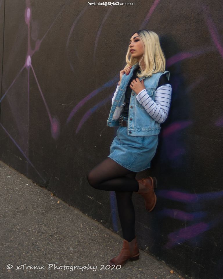 A person with blonde hair wearing a denim jacket and a striped shirt stands against a wall with colorful graffiti, striking a casual pose.